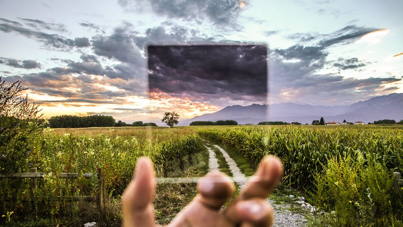 Una mano sostiene un filtro de cristal sobre un camino entre campos al atardecer; a través del cristal, el mismo cielo se ve distinto