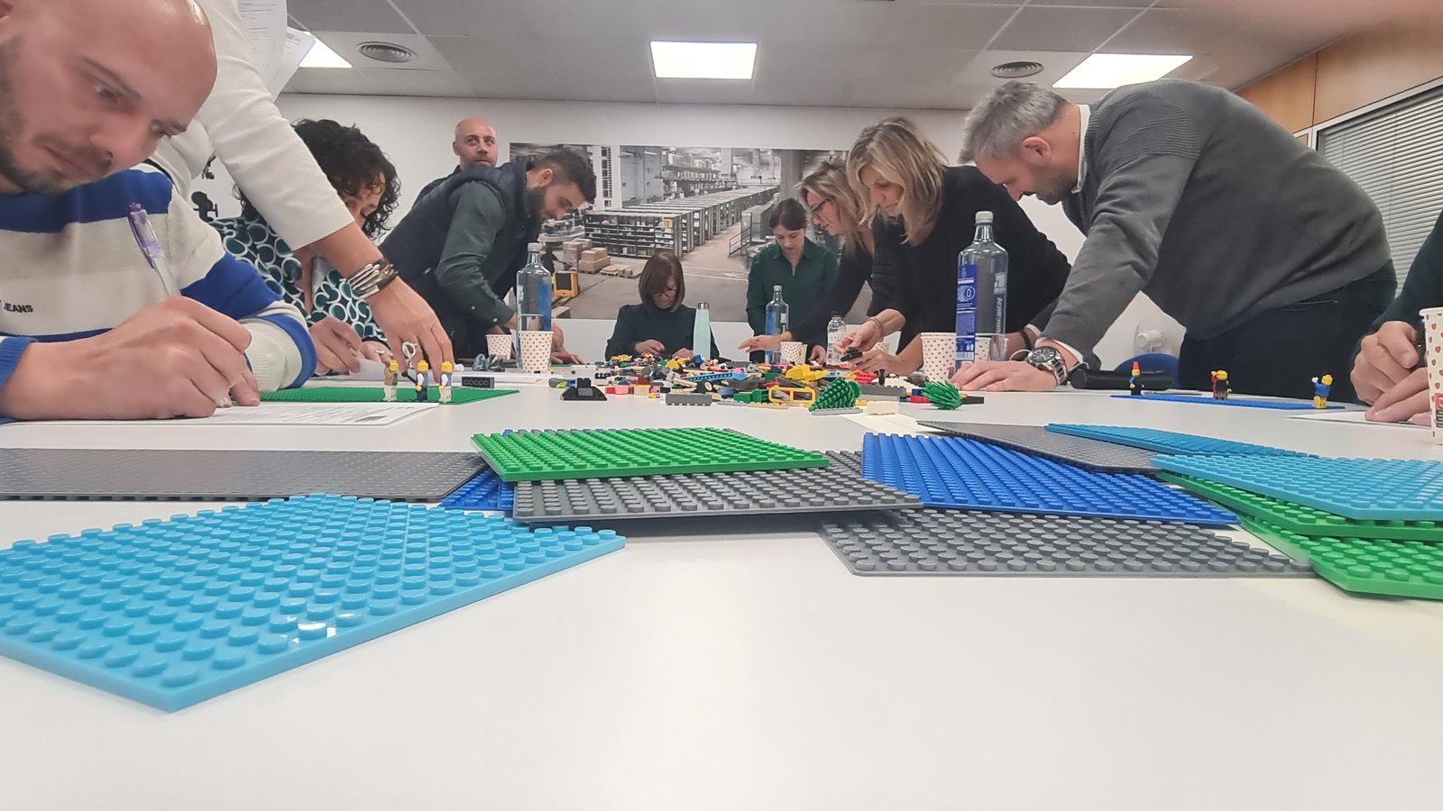 Equipo directivo de pie alrededor de una mesa, construyendo con LEGO sobre placas base de colores
